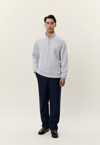 Giovane uomo in piedi che indossa una felpa grigio chiaro con zip a quarto, con scritta "LD", pantaloni blu navy e scarpe di pelle nere, sfondo neutro.
