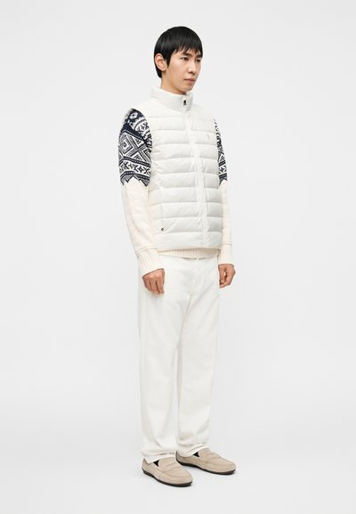 Λευκό αμάνικο γιλέκο με μπλε σκούρα μανίκια με σχέδιο, φορεμένο πάνω από κρεμ πουλόβερ και λευκά παντελόνια. Αξεσουάρ περιλαμβάνουν ανοιχτό μπεζ παπούτσια χωρίς δέσιμο.