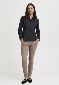 Camicia nera a maniche lunghe, con colletto e bottoni, abbinata a pantaloni beige attillati e mocassini neri con dettagli dorati.