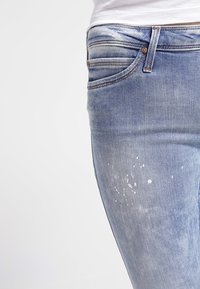 Jeans de denim azul claro con un lavado desgastado y salpicaduras de pintura blanca en el muslo, usados con una camisa blanca metida por dentro.