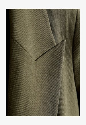 Olíva zöld, texturált szövet, szabott kialakítással, szöges varrási részletekkel és strukturált sziluettel.