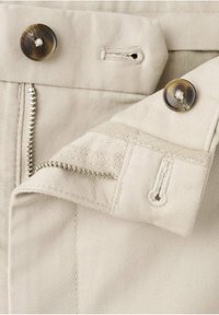 Pantalon beige avec des boutons en écaille de tortue, des boutonnières visibles et une fermeture éclair métallique partiellement ouverte sur un tissu texturé.