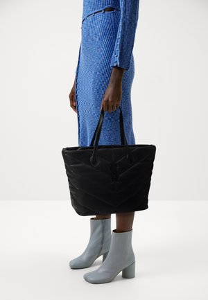 Schwarze, gesteppte Tragetasche mit langen Griffen, die ein strukturiertes Chevron-Muster aufweist. Die Tasche wird von einer Person in einem blauen Rippenkleid und hellblauen Stiefeln gehalten.