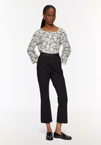 Un pantalon noir taille haute associé à un blouse à manches longues à motif featuring des lignes noires ondulées sur fond clair, complété par des mocassins noirs.