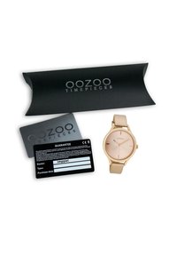 Montre-bracelet dorée avec un cadran simple, un bracelet beige et un design minimaliste, accompagnée d'une boîte noire et d'une carte de garantie.