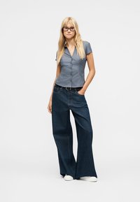 Mujer rubia con gafas, camisa de manga corta azul grisácea abotonada, vaqueros de pierna ancha oscuros y zapatos blancos, de pie con una mano en el bolsillo.