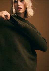 Dunkelgrüner Pullover aus einem weichen, strukturierten Stoff, mit Rundhalsausschnitt und langen Ärmeln. Ein auffälliges Detail ist die übergroße Öffnung am Ärmelbund.