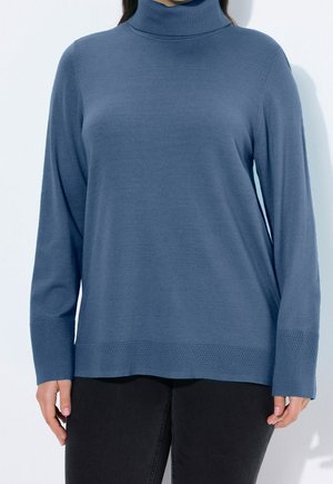 Personne portant un pull à col roulé bleu avec des poignets et un ourlet texturés, associé à un pantalon noir sur un fond clair uni.