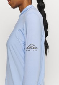 Helles blaues Langarmshirt aus glattem Material mit einem funkelnden schwarzen "Nike Trail"-Logo auf dem linken Arm.