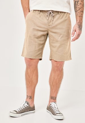 Beige cotton shorts with an elastic waistband and drawstring, featuring side pockets and a relaxed fit. Worn with gray canvas sneakers.