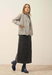 Cardigan gris de punto con bolsillos de parche sobre unsuéter de cuello alto gris, combinado con una falda larga de mezclilla negra y botas de tacón azul brillantes.