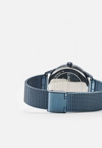 Montre en acier inoxydable bleue avec un bracelet en maille, possédant un boîtier rond et un fermoir gravé, mettant en avant un design minimaliste et un dos argenté.