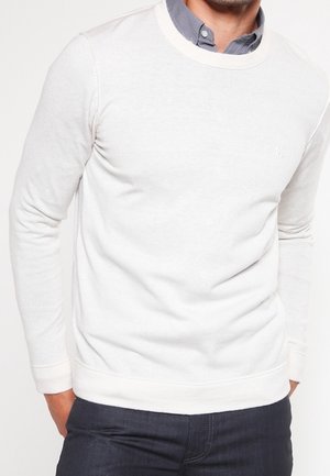 Weißer Pullover mit Rundhalsausschnitt und langen Ärmeln, mit einer strukturierten Strickoberfläche und einem gerippten Saum, über einem Kragenhemd getragen.