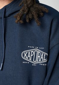 Sweat-shirt bleu marine avec un logo frontal lisant "KAPORAL" en blanc. Il dispose d'une capuche avec cordon de serrage et d'un tissu doux et texturé.