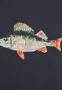 Diseño de pez bordado en una tela azul marino oscuro, con acentos verdes y rojos y detalles texturizados en las escamas y aletas del pez.