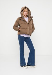 Casaco puffer castanho com capuz, textura acolchoada, fecho frontal, usado sobre uma blusa lilás clara e jeans de boca de sino azuis, com sapatos pretos.