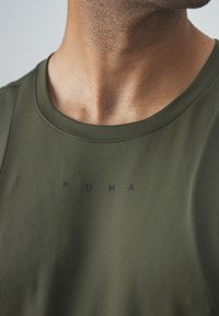 Primer plano del cuello y parte superior del pecho de un hombre que lleva una camiseta sin mangas de Puma en verde oliva con letras del logo en negro centradas en la tela.