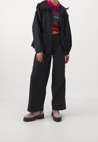 Chaqueta ligera negra con capucha y cintura ceñida. Debajo, una camiseta corta de rayas multicolores. Pantalones negros de pierna ancha y zapatos rosas con cordones.
