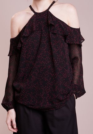 Kobieta ubrana w czarną przezroczystą bluzkę z odkrytymi ramionami, ozdobioną bordowymi gwiazdkami i falbanami, w połączeniu z czarnymi spodniami.