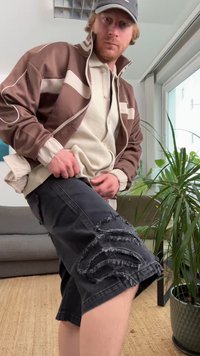 Brązowa sportowa kurtka z białymi akcentami na kremowej, fakturowanej koszuli, w połączeniu z czarnymi, postrzępionymi szortami z ozdobnymi przebarwieniami.