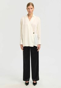 Chemise en tissu beige clair à manches longues et col, dotée d'une patte sur le côté et d'un détail de bouton argenté, associée à un pantalon noir à jambe large.