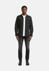 Camisa negra abotonada con dos bolsillos en el pecho, llevada sobre una camiseta blanca. Combinada con jeans gris oscuro y zapatos negros. Diseño simple.