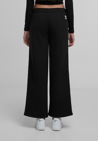 Črne široke trenirke z teksturirano površino, elastičnim pasom in majhnim logotipom na zadnjem žepu. V kombinaciji z belimi športnimi copati.