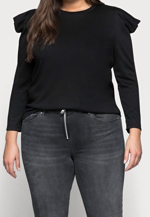 Kvinde iført sort langærmet top med flæseærmer og falmede sorte jeans med høj talje og synlig lynlås foran.