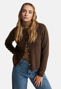 Hnědý pletený kardigan s kulatým výstřihem a perlami, přes černý a béžový pruhovaný rolák, kombinovaný s modrými džíny.