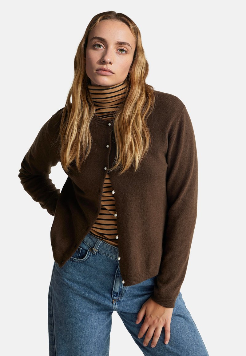 Hnědý pletený kardigan s kulatým výstřihem a perlami, přes černý a béžový pruhovaný rolák, kombinovaný s modrými džíny.