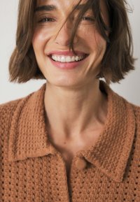 Brown knitted cardigan with a textured, open stitch design. Features a collar and round button, showcasing a warm, earthy color palette.