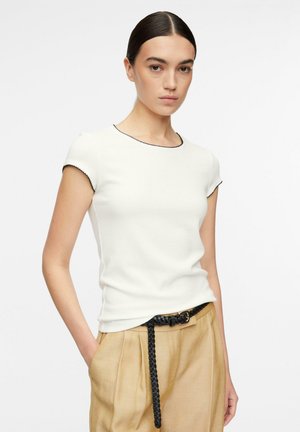 Woman with dark slicked-back hair wears white fitted short-sleeve top with black trim and beige high-waist pants with a braided black belt.