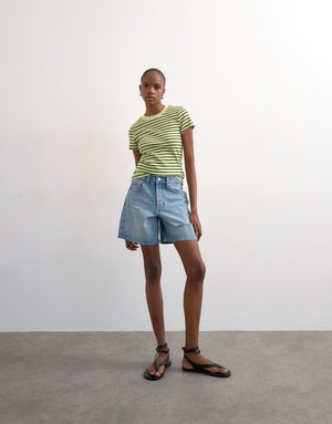 Frau trägt ein hellgrünes und schwarz gestreiftes T-Shirt, lockere Jeans-Shorts und schwarze Sandalen mit Riemchen, steht auf einem grauen Boden vor einer weißen Wand.