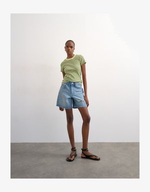 Frau trägt ein hellgrünes und schwarz gestreiftes T-Shirt, lockere Jeans-Shorts und schwarze Sandalen mit Riemchen, steht auf einem grauen Boden vor einer weißen Wand.