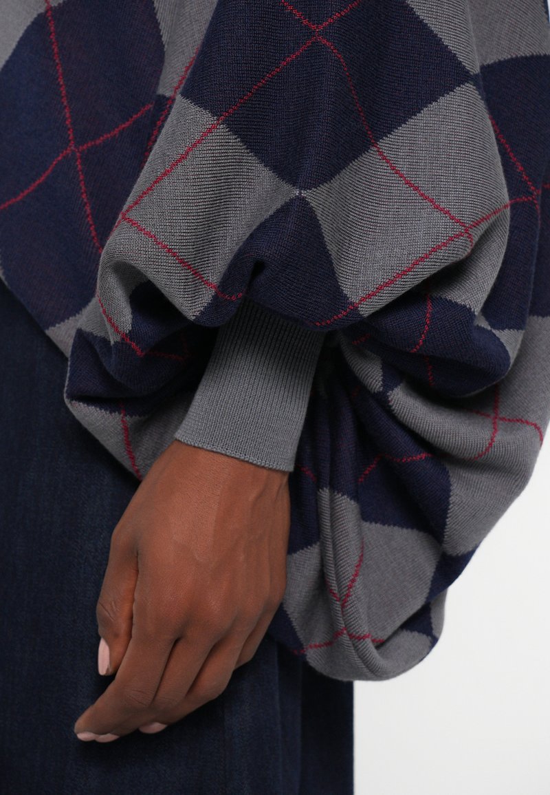 Maglione grigio e blu navy con motivo argyle e accenti rossi; presenta una manica larga e voluminosa con polsini a costine. Mano parzialmente visibile.