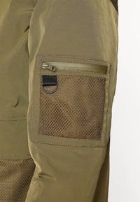 Detailní záběr na kapsu zelených cargo kalhot s mesh panely, zipovým uzávěrem a černým textilním poutkem s kovovým kroužkem.