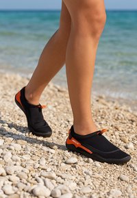 Chaussures d'eau noires avec des accents orange, fabriquées en matériau flexible, dotées d'un design à enfiler, de semelles texturées et d'un ajustement sécurisé.