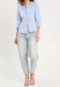 Femme portant un chemisier rayé bleu clair et blanc avec des liens, un jean taille haute délavé clair et des sandales dorées à bout ouvert, debout sur un fond uni.