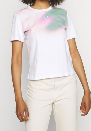 Personne portant une chemise blanche à manches courtes avec des motifs éclaboussés rose pastel et vert près de l'encolure et un pantalon beige.