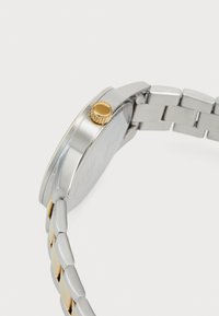 Sølv armbåndsur med et tofarvet metalrem og en guldkronen på siden, fotograferet fra en tæt vinkel fra siden.