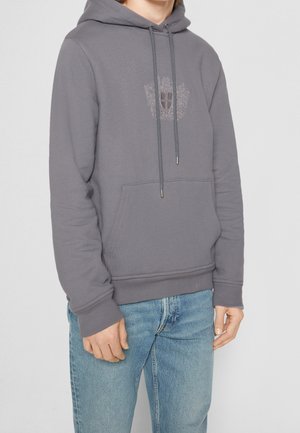 Mand iført grå hættetrøje med forlommer og broderet våbenskjold, kombineret med lyseblå jeans, stående mod en ensfarvet baggrund.