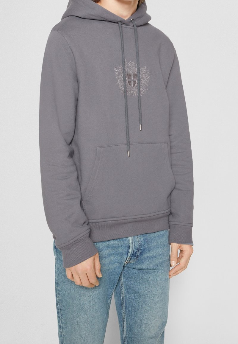 Homme portant un sweat à capuche gris avec poche avant et blason brodé, associé à un jean bleu clair, debout devant un fond uni.