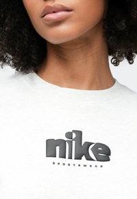 Κοντινό πλάνο ενός ατόμου που φοράει λευκό πουκάμισο με έντονο μαύρο λογότυπο "nike SPORTSWEAR" στο στήθος.