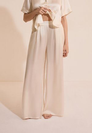 Person trägt locker sitzende, cremefarbene, weit geschnittene Hose mit hoher Taille und passendes kurzärmeliges Hemd mit Knöpfen, hebt das Hemd leicht an.