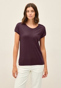 Kortärmad, rundhalsad lila t-shirt i mjukt tyg med plisserade axeldetaljer, tillsammans med vita byxor.