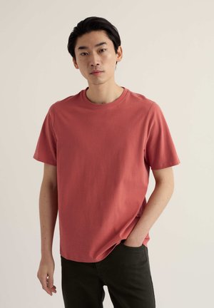 Kurzärmeliges T-Shirt mit Rundhalsausschnitt in verblasstem Korallton. Hergestellt aus Baumwolle mit glatter Textur, kombiniert mit dunkler Hose.