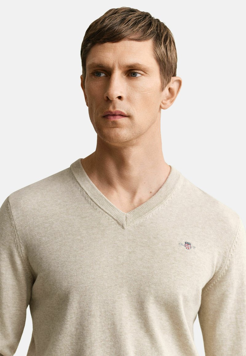 Mann mit kurzen braunen Haaren, der einen beigefarbenen V-Ausschnitt-Pullover mit einem kleinen bestickten Wappen auf der Brust trägt und zur Seite schaut.