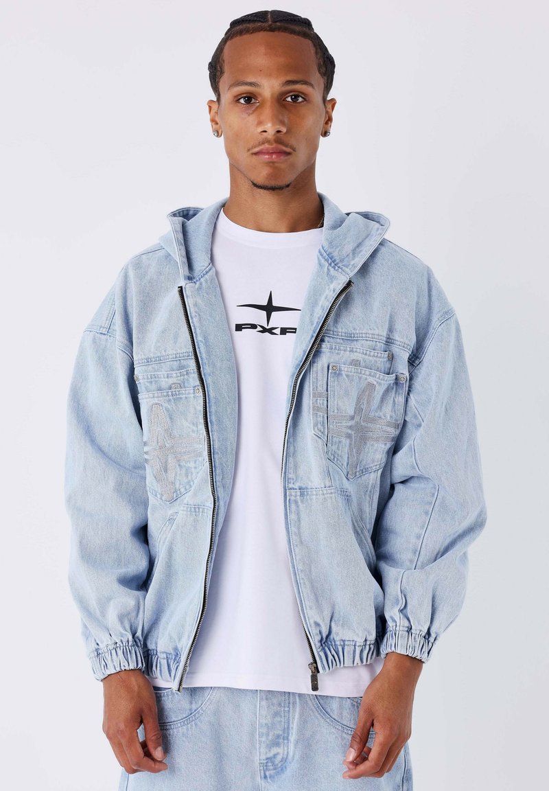Veste en denim bleu clair avec fermeture éclair à l'avant, poignets élastiques et deux poches avant avec un design texturé. Portée sur un T-shirt blanc.