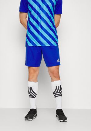 Mężczyzna ubrany w niebieską sportową koszulkę w paski, niebieskie spodenki Adidas, białe wysokie skarpetki z czarnymi wzorami oraz czarne korki stoi przy białej ścianie.