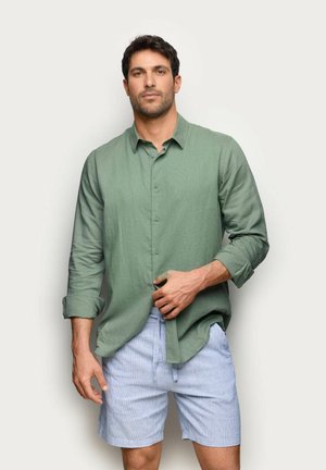 Homme aux cheveux courts et foncés portant une chemise verte boutonnée et un short bleu clair, debout devant un fond clair uni.